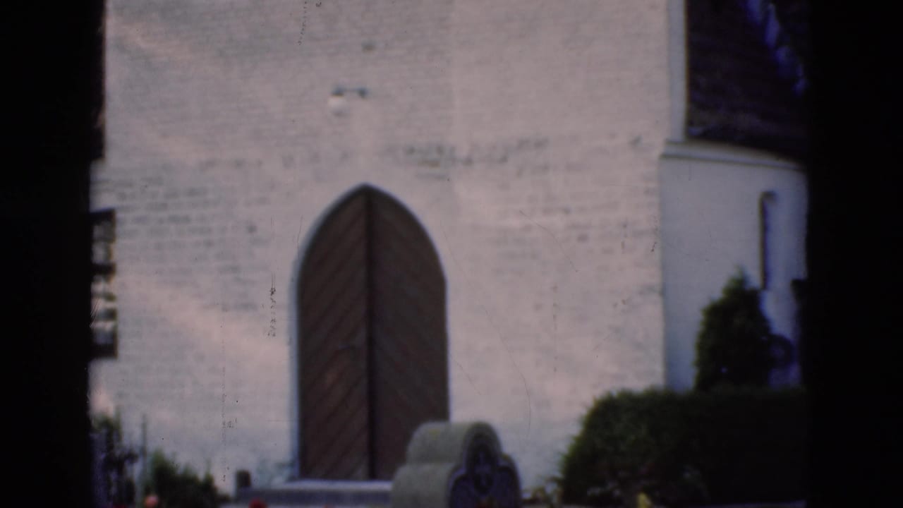 1975 Denmark 8mm view of a whitewashed church entrance framed by cemetery stones, shrubs, and a dark foreground tree.