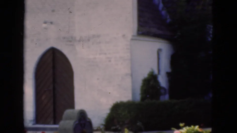 1975 Denmark 8mm frame with the arched church door, nearby gravestone, clipped hedge, and shaded planting at the churchyard edge.
