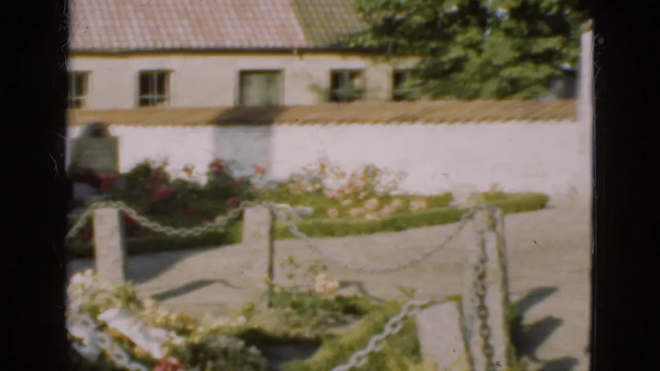 1975 Danish cemetery view showing chain edging, planted graves, a white wall, and a tiled-roof building beyond.