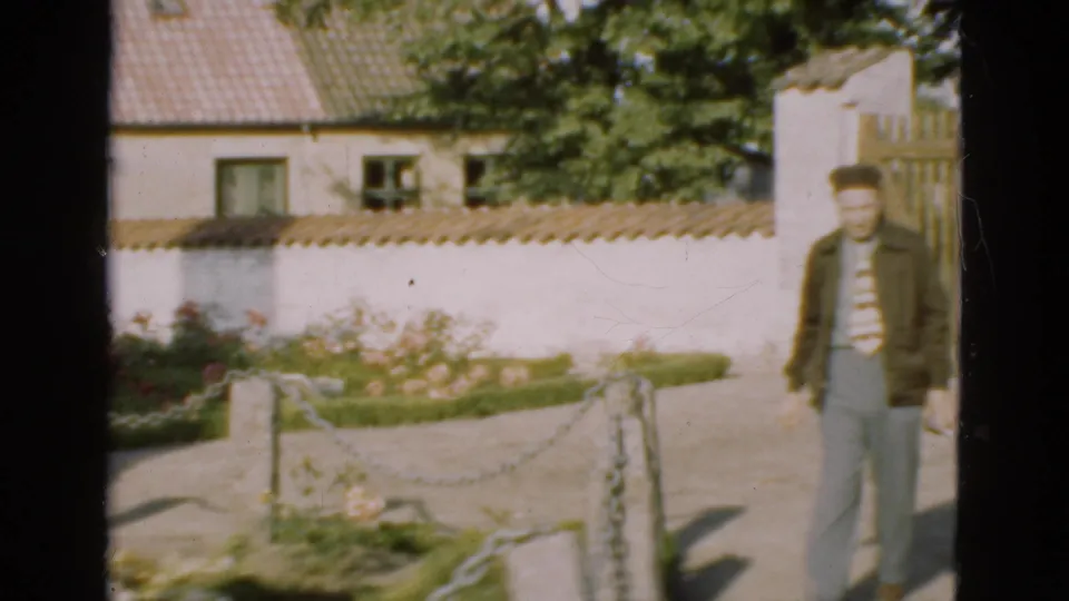 1975 Danish 8mm frame with an adult man walking through cemetery grounds near flowers, chain edging, and a white wall.