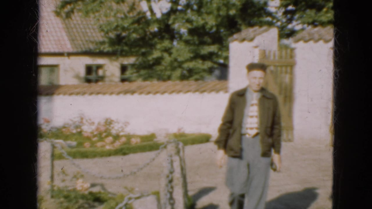 In 1975 Denmark, an adult man walks along a sunny garden path beside a whitewashed wall and wooden gate.