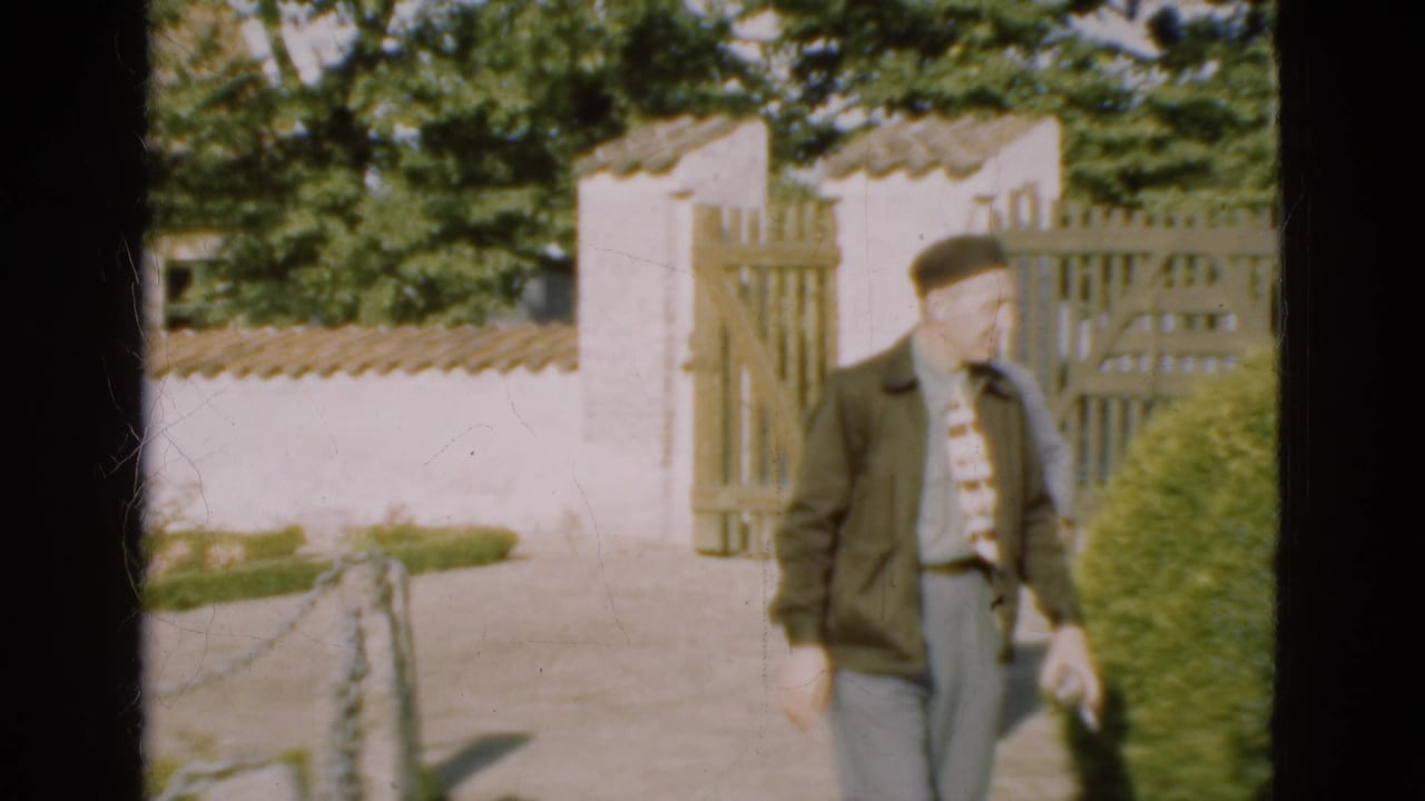 In 1975 Denmark, a man in a dark jacket and tie walks beside a hedge near a rural wooden garden gate.