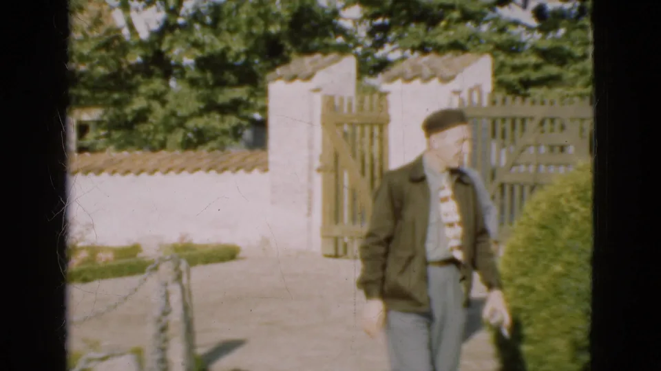 In 1975 Denmark, a man in a dark jacket and tie walks beside a hedge near a rural wooden garden gate.