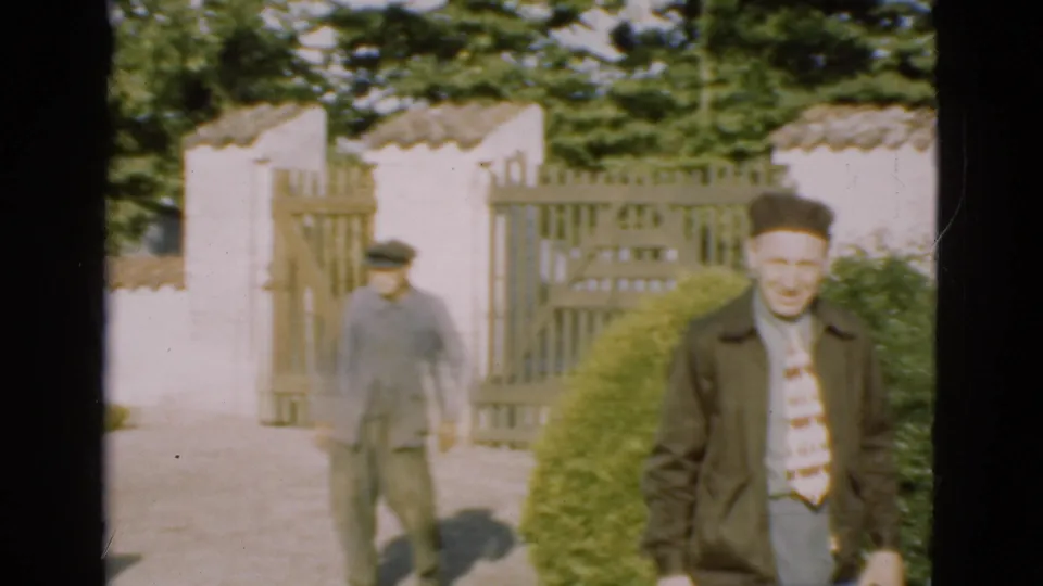 In 1975 Denmark, two adult men walk through a rural garden entrance, with one man close to the camera and another behind him.