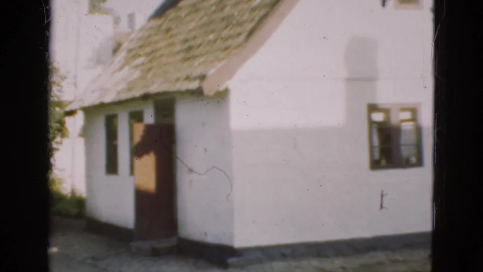 In 1975 Denmark, a whitewashed rural house with a thatched roof and open doorway appears in bright daylight.