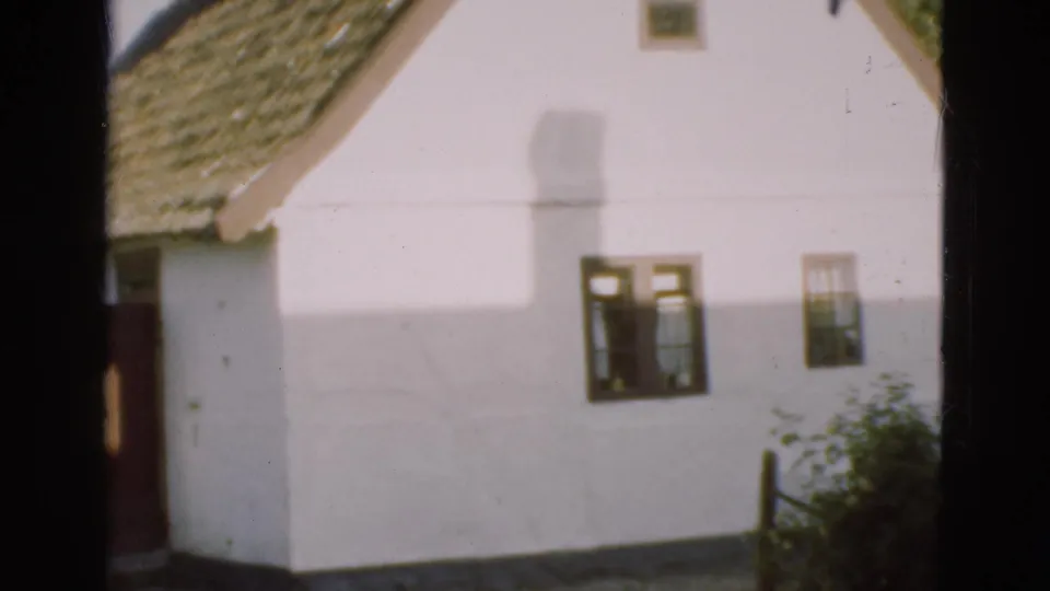 1975 Denmark 8mm frame showing the side wall of a white thatched-roof house with small windows, garden greenery, and a fence post.