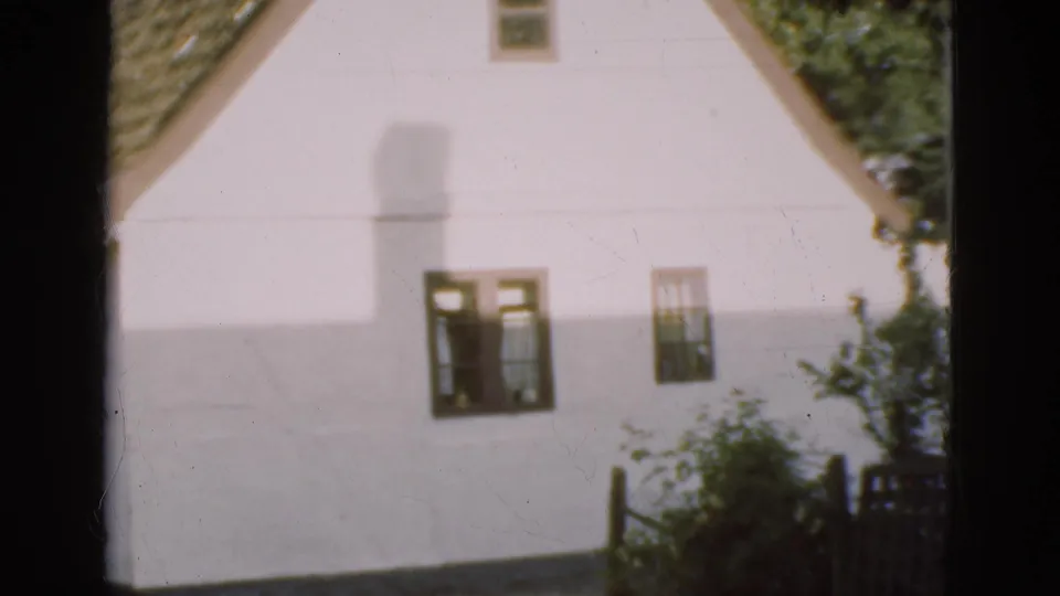 1975 Denmark home-movie frame of a white house wall with small dark-framed windows, roof edge, garden shrubs, and a fence at the right.