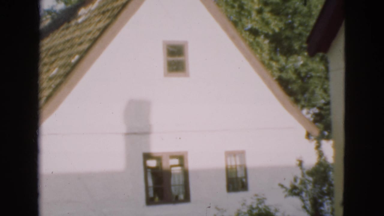 1975 Denmark 8mm frame of a white house with a steep triangular gable, small windows, roof tiles and trees in the background.