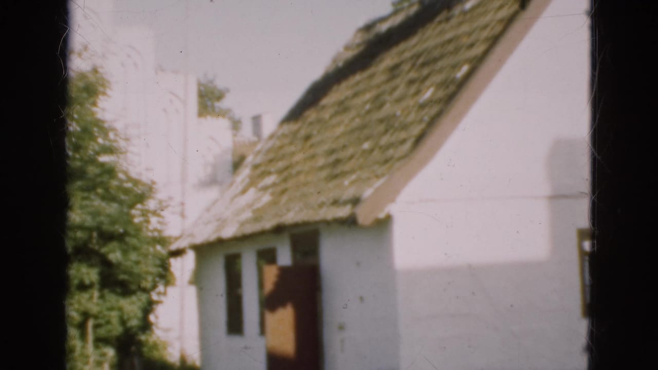 1975 Denmark exterior frame with the camera closer to the white thatched building, showing its doorway, roof, and nearby trees.