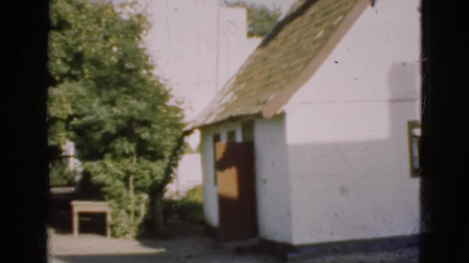1975 Denmark 8mm frame showing the side wall of a white thatched building, leafy trees, and a small bench-like object near the ground.