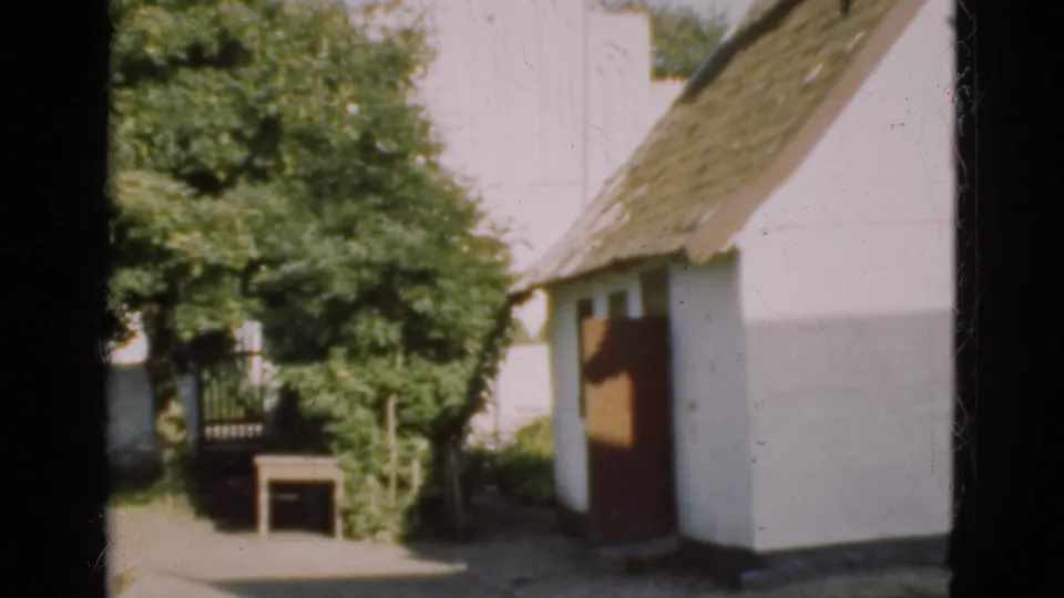 1975 Denmark home-movie frame showing a white thatched-roof building with a dark doorway, trees, and bright outdoor light.