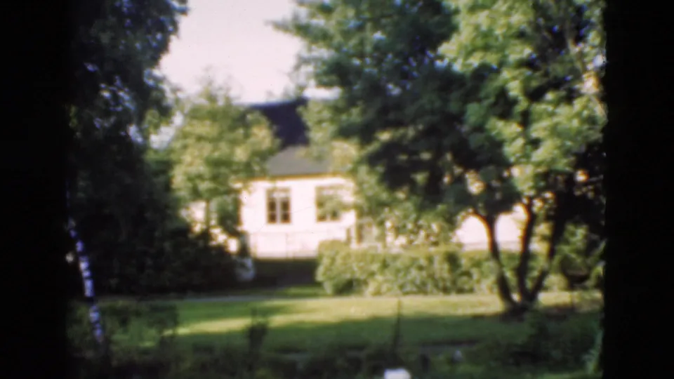 1975 Denmark 8mm frame of a sunlit garden, trees, shrubs, and a light-colored house in the background.