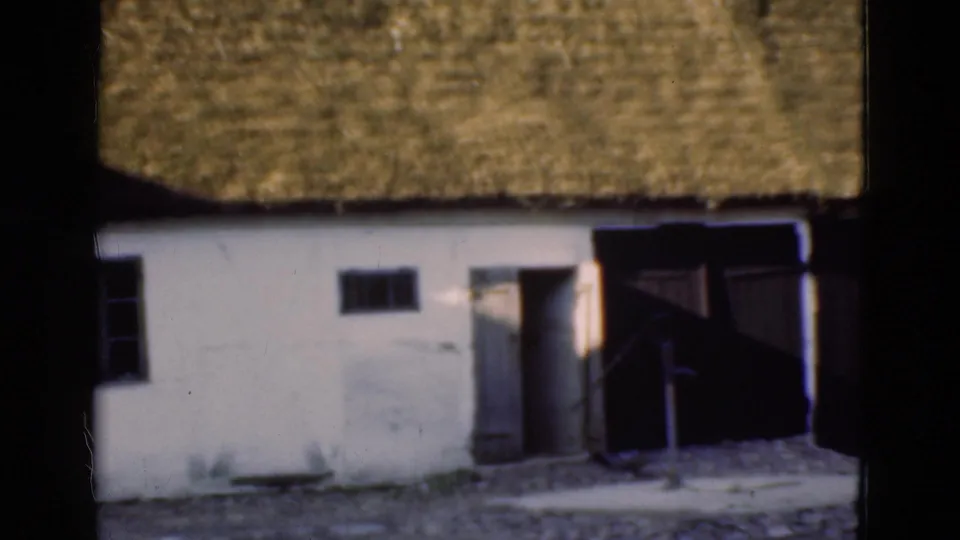 1975 Denmark 8mm frame with a white thatched-roof building, small windows, open doorway, and dark wooden doors to the side.