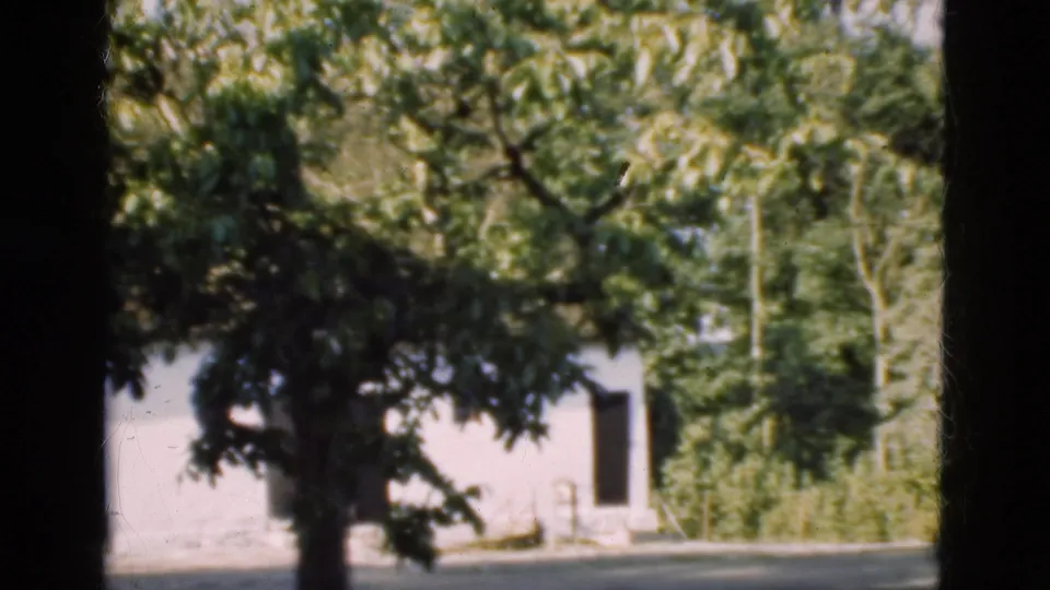 1975 Denmark frame showing a leafy tree before a white rural building with a dark open doorway and yard.