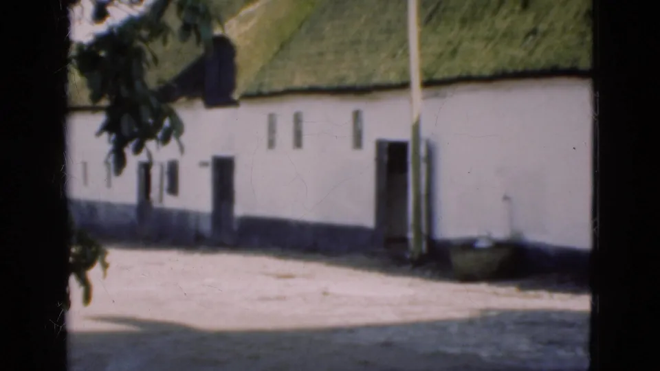 1975 Denmark 8mm frame showing a white thatched-roof rural building with dark openings and a dirt yard.