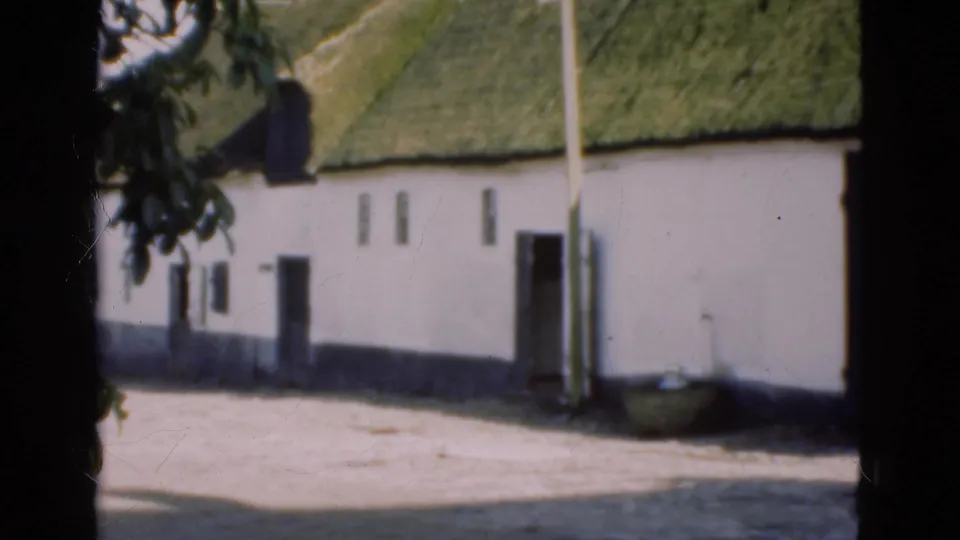 1975 Denmark 8mm frame showing a whitewashed building side with moss-green thatched roof, small openings, doorway, pole, and pale yard ground.