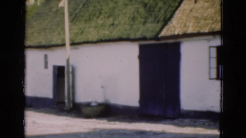 1975 Denmark 8mm frame showing the building side as the pan reaches a dark wooden door, with thatch, pole, basin, and a small window visible.