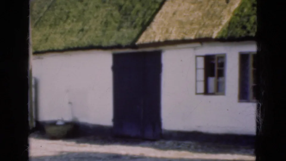 1975 Denmark frame showing a large dark door on a whitewashed thatched building, with an open window, basin, low base, and yard foreground.