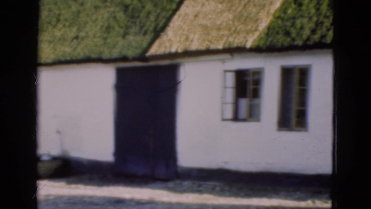 1975 Denmark 8mm frame showing part of a white cottage with a green thatched roof, dark door, and wooden-framed windows.