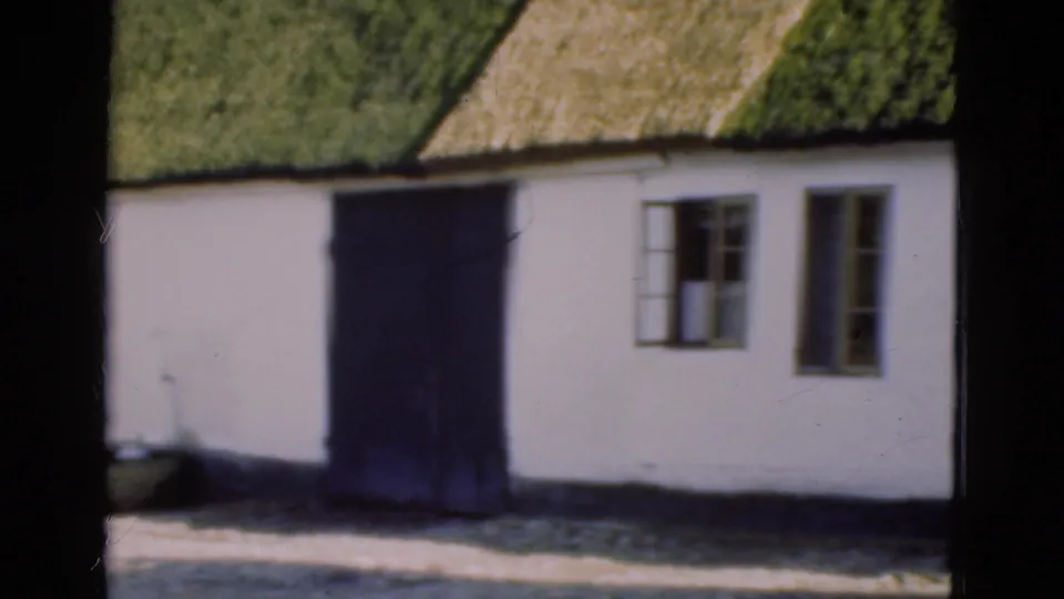 1975 Denmark 8mm frame showing part of a white cottage with a green thatched roof, dark door, and wooden-framed windows.