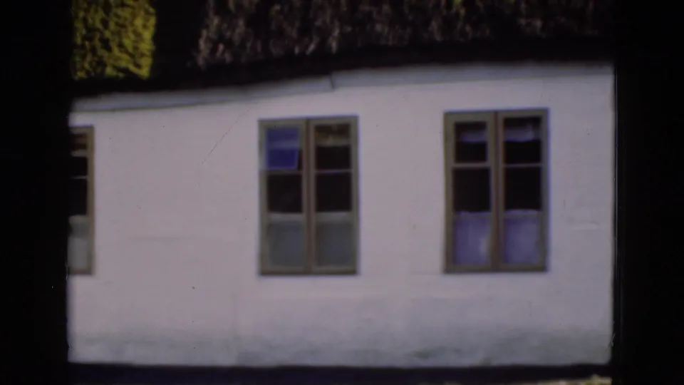 1975 Danish 8mm frame showing two main windows in a white wall below a dark thatched roof, with curtains visible inside.
