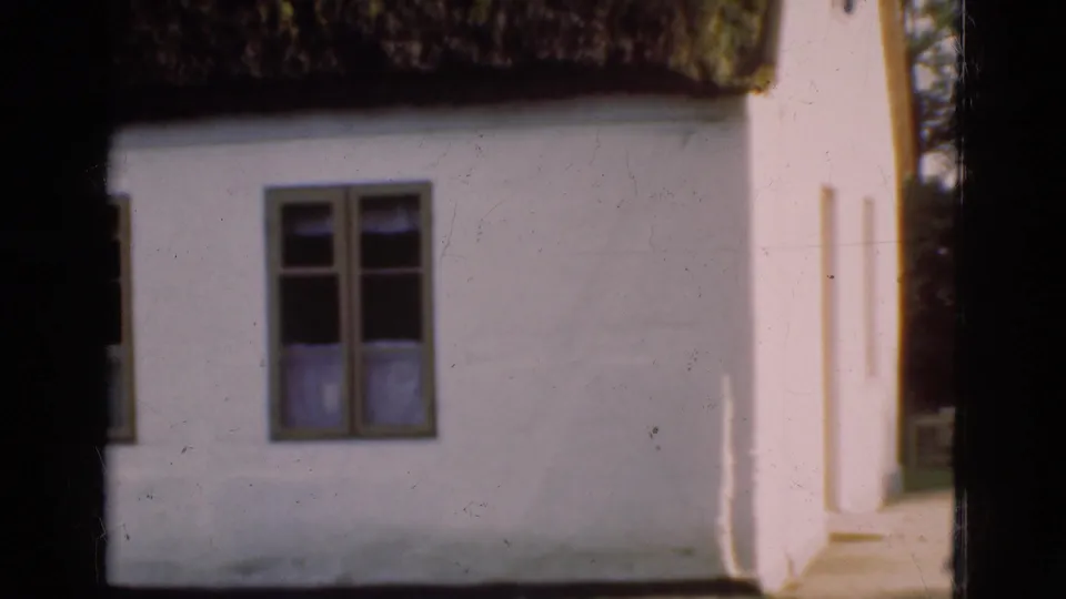1975 Danish 8mm frame showing the building corner, a single multi-pane window, thatched roof edge, and a bright side passage.