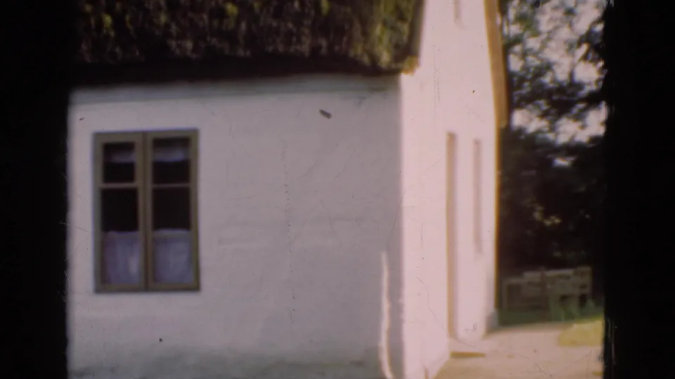 1975 Denmark 8mm frame showing a whitewashed cottage facade, small window, curtains, and thick thatched roof edge.