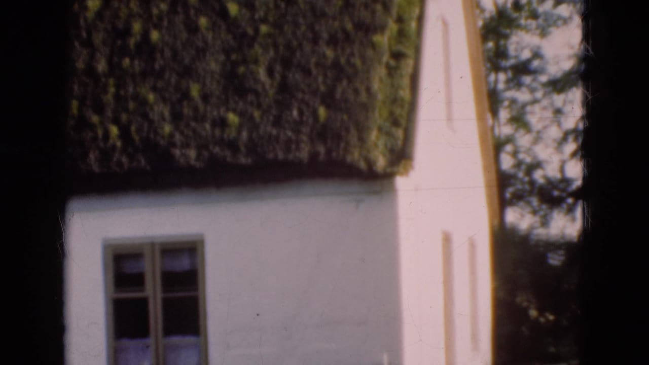 1975 Denmark home-movie frame of a thatched cottage wall and window, partly framed by dark scan borders and nearby trees.