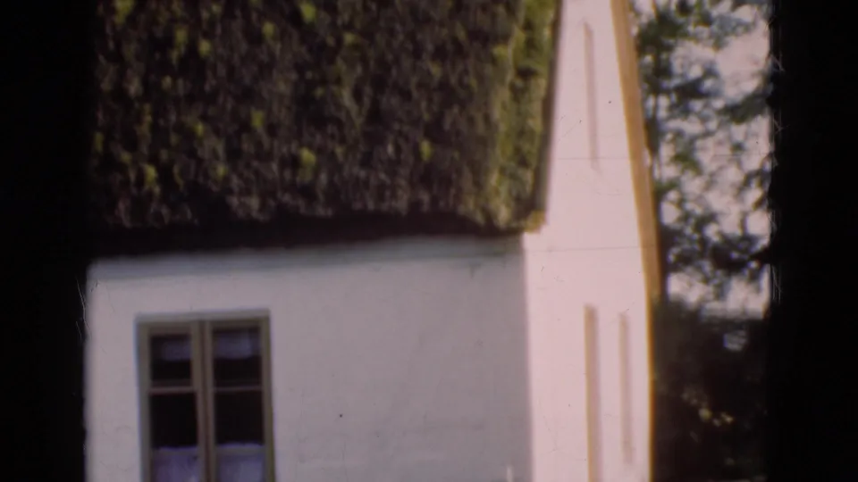 1975 Denmark home-movie frame of a thatched cottage wall and window, partly framed by dark scan borders and nearby trees.