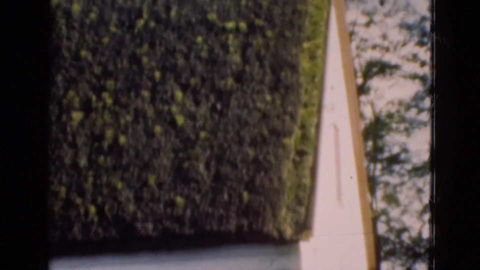 1975 Denmark 8mm frame focusing on the heavy thatched roof edge, white wall, and shaded trees beside the cottage.