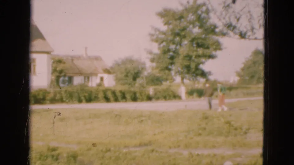 1975 Denmark 8mm frame showing a grassy yard, thatched cottages, trees, hedges, and small distant figures walking near a path.