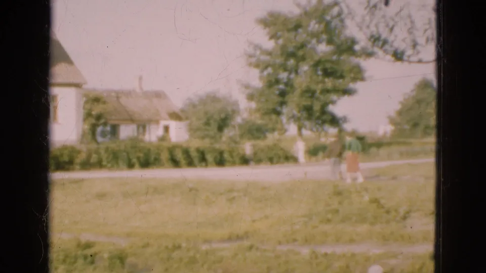 1975 Denmark 8mm frame showing a distant person in a green field, roadside houses, trees, and a dark window frame in the foreground.