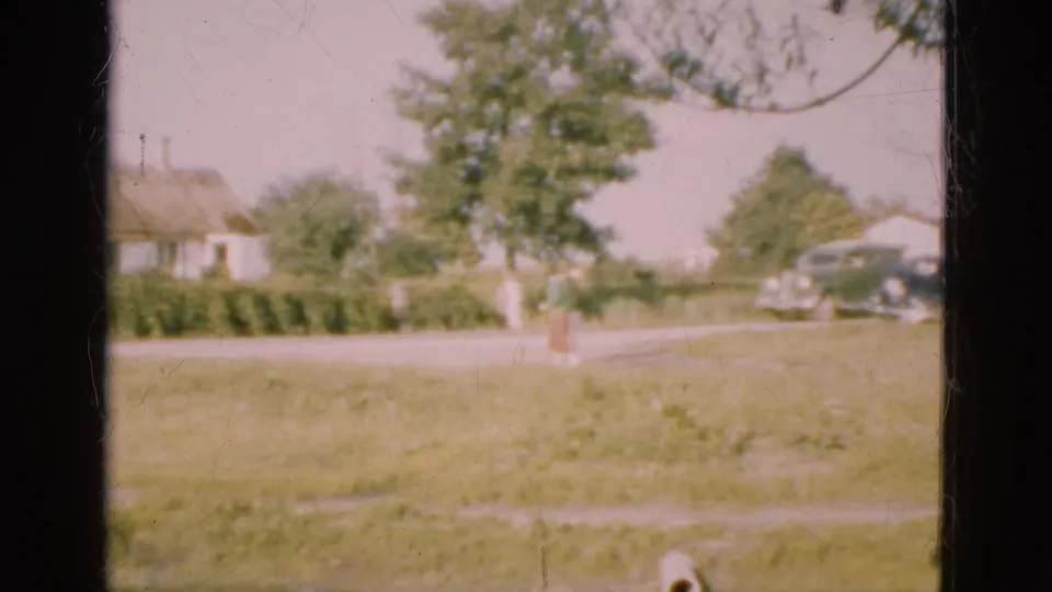 1975 Denmark home-movie frame with a person near a tree, a grassy field, roadside houses, and cars visible beyond the window frame.