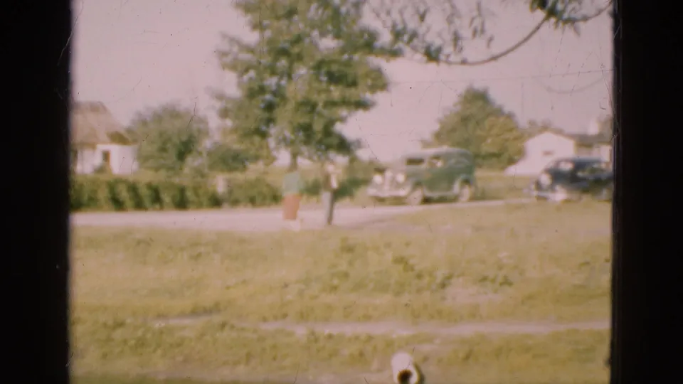 Soft 8mm frame from Denmark in 1975 showing a grassy field, a person near a tree, roadside houses, and classic cars in the distance.