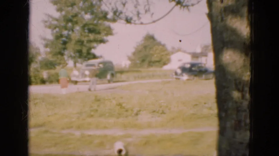 1975 Denmark 8mm frame showing a person near a tree, two cars by the road, low houses, grass, and a dark window frame at the edge.