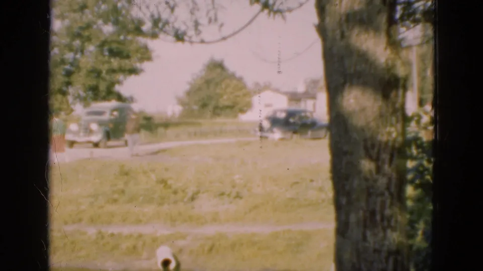 1975 Denmark home-movie frame looking through a window at a small road, cars, lawn, trees, and houses beyond.