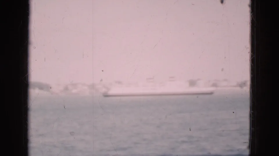 Soft 8mm footage from Denmark in 1975 frames a ship on harbor water with low shoreline buildings beyond.