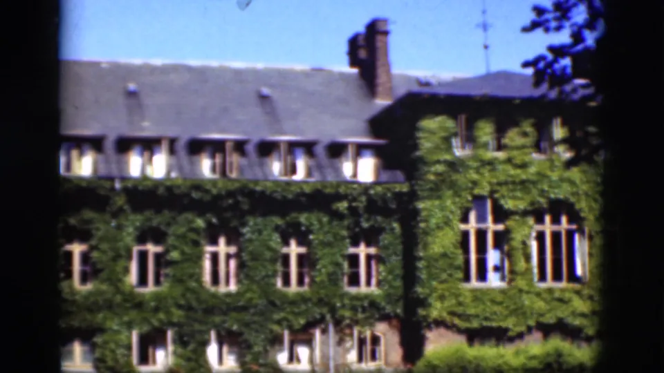 1975 Denmark 8mm frame of the ivy-covered house, showing the roofline, brick chimneys, repeated windows, foreground foliage, and film-edge obstruction.