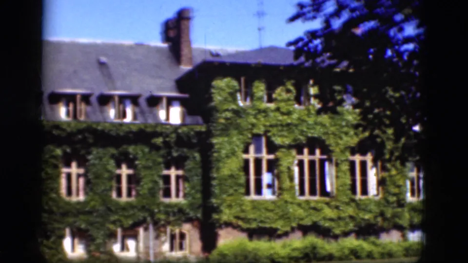 1975 Denmark 8mm frame showing a closer exterior view of the vine-covered house, with tall windows, roof dormers, shrubs, and branches at the edge.