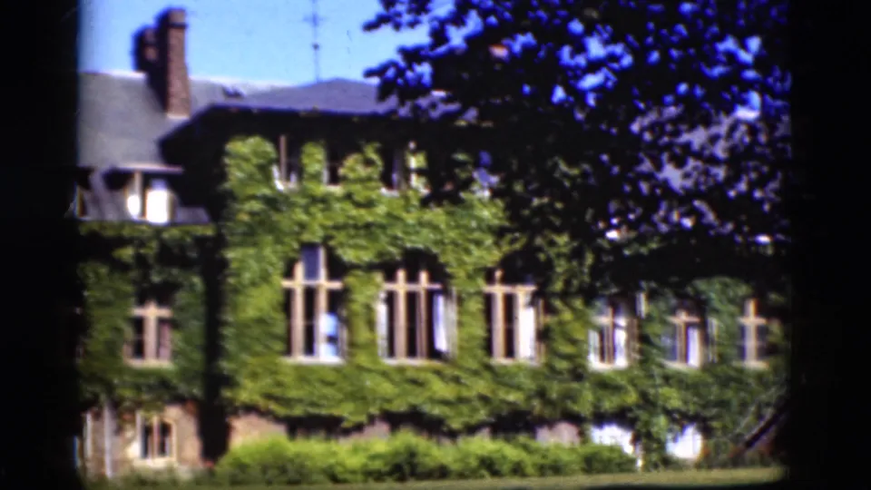 1975 Denmark 8mm frame of the ivy-covered building partly obscured by tree branches, with tall windows, shrubs, dark roof, and blue sky visible.