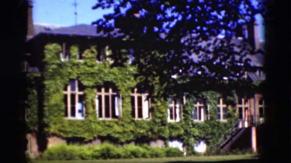 1975 Denmark home-movie frame showing an ivy-covered building facade, tall windows, shrubs, lawn, and dark tree branches overhead.