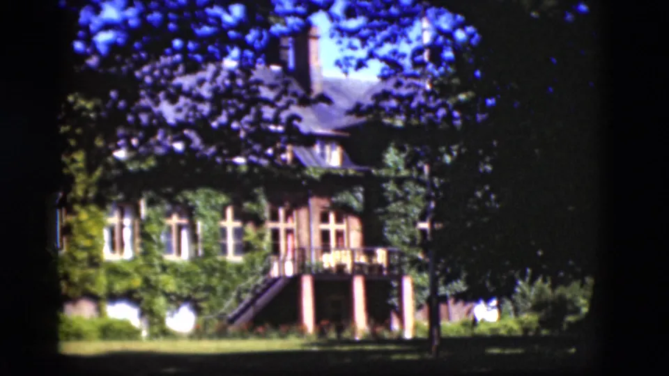 1975 Denmark home-movie view of the ivy-covered building, exterior stairs, lawn, and a dark tree trunk crossing the foreground.