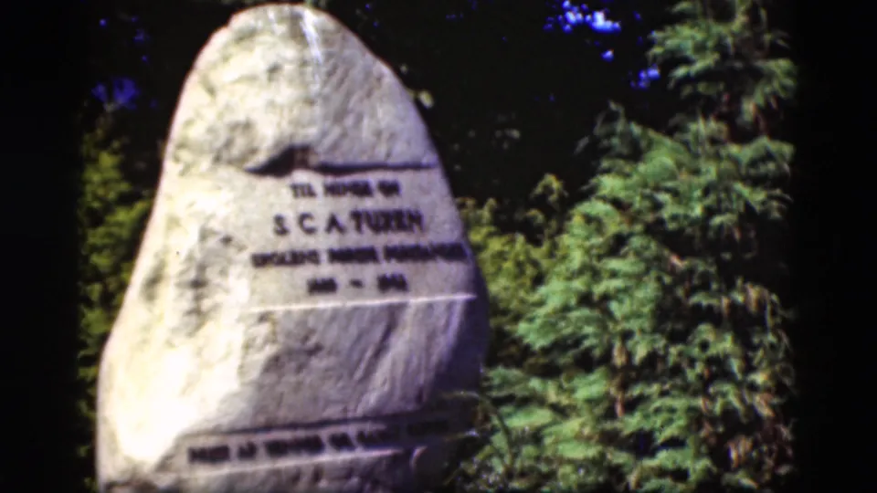 1975 Denmark frame showing a pale memorial stone with partially legible lettering, surrounded by shrubs and evergreen trees.