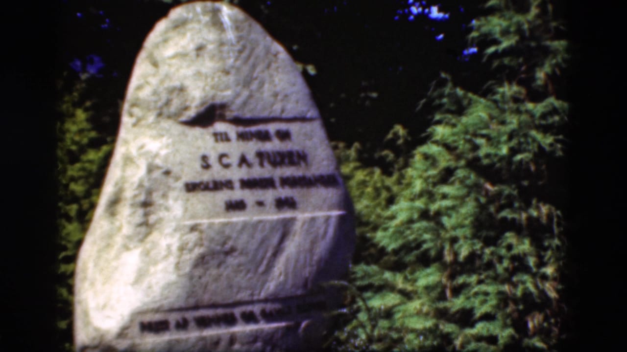 1975 Denmark 8mm frame showing a tall stone memorial with Danish inscription, surrounded by dark trees and greenery.