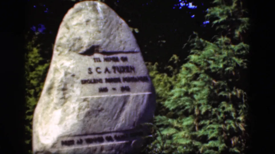1975 Denmark 8mm frame showing a tall stone memorial with Danish inscription, surrounded by dark trees and greenery.