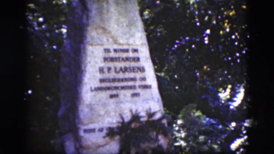 1975 Denmark 8mm frame focused on the H.P. Larsen memorial stone in a shaded, leafy cemetery setting.