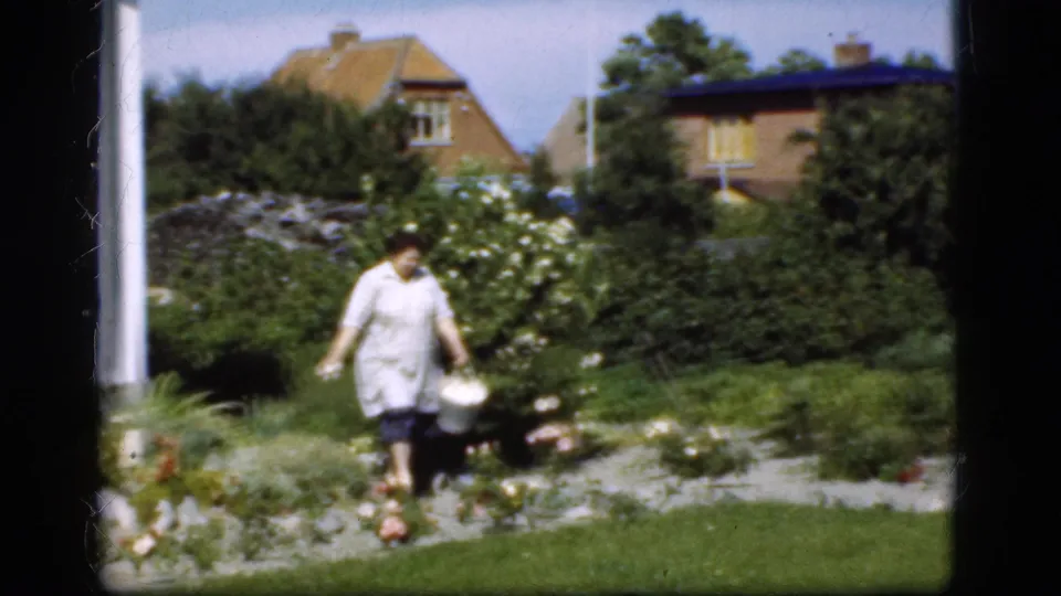 In Denmark 1975 home-movie footage, a woman in a light dress carries a small white pail through a garden with flowers and shrubs.