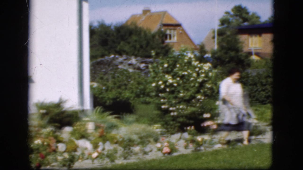 Woman with a pail continues walking across the garden in front of shrubs and houses.