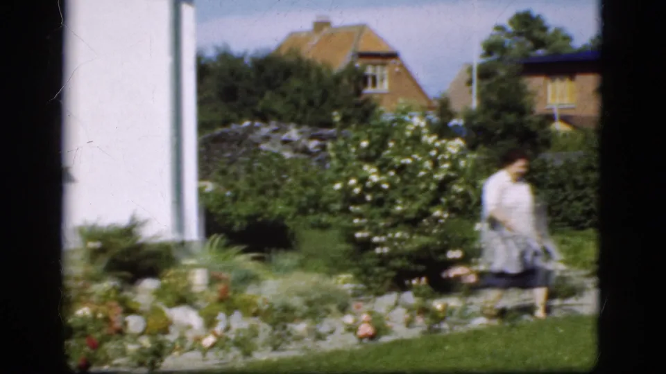 The woman walks farther across the Danish garden, with flowering shrubs, lawn, and neighboring house roofs visible behind her.