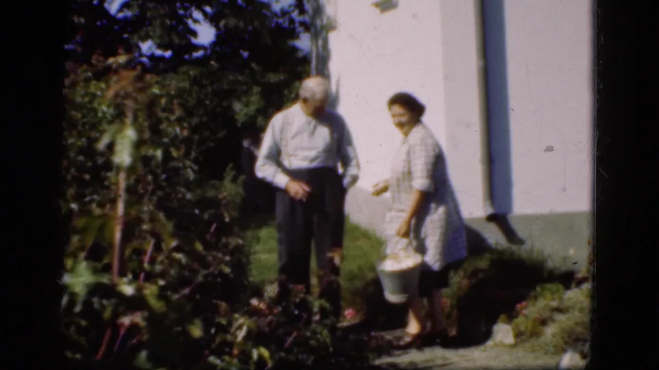 1975 Denmark 8mm frame of an older man and woman in a garden beside a white house, with the woman holding a bucket.